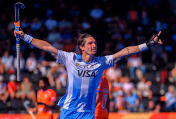 Un jugador de hockey con camiseta celeste y blanca celebra con los brazos extendidos y lleva un palo en la mano